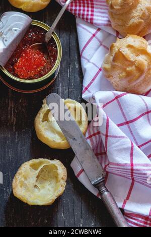 Home Made Profiteroles geschnitten und fertig zum Zubereiten für roten Kaviar mit Vintage-Messer und Dose roten Kaviar auf weißem Küchentuch über Holztisch. Dunkles r Stockfoto