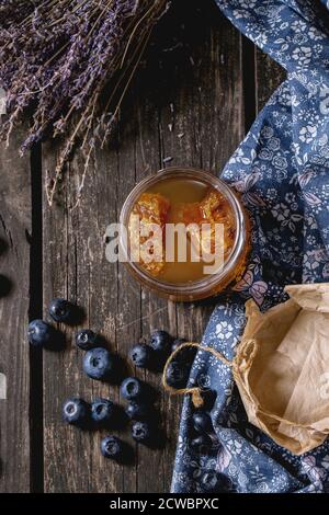 Glas Glas von flüssigen Honig mit Wabenstruktur innen, frische Blaubeeren und Bündel trockener Lavendel über alten Holztisch mit blauen Textilien rag Öffnen. Dunkle ru Stockfoto