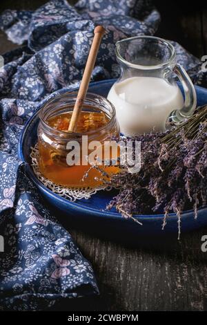 Offenes Glas mit flüssigem Honig mit Honigwabe und Honig-Dipper innen, Glaskrug mit Milch und Bund von trockenem Lavendel in blauer Keramikplatte über Holz s Stockfoto
