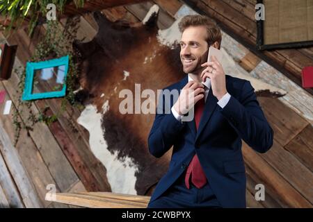 Junger Geschäftsmann, der am Telefon spricht, lächelt und Krawatte festmacht und in einem Coffeeshop zur Seite schaut Stockfoto