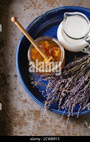 Öffnen Glas Glas von flüssigen Honig mit Wabe und Honig Pendelarm innen, Glas kanne Milch und Bündel trockener Lavendel in großen blauen Keramik Platte über alte Stockfoto