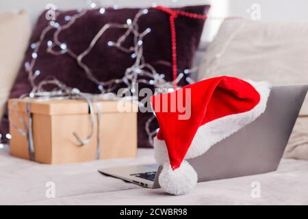 Weihnachtslaptop in weihnachtsmütze auf einem Sofa stehend, ein freier Platz. Remote Work am Heiligabend. Online-Shop, Online-Bestellung Geschenke für die Feiertage. Stockfoto