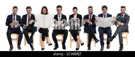 Team von 8 Geschäftsleuten lesen von Tablet, Telefon, Buch und Zeitung, während auf Stühlen auf weißem Studio Hintergrund sitzen Stockfoto