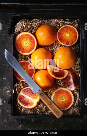 Scheiben und ganze sizilianische Blut Orangen Obst in schwarzer Holzkiste mit Sägemehl auf dunklem Hintergrund. Mit Vintage-Messer. Flach liegend Stockfoto