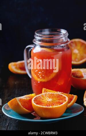 Sizilianische Blutorangen in Scheiben geschnitten in türkisfarbenen Keramikplatte und Glas-Einmachglas von frischen roten Orangensaft über alten Holztisch. Dunkler rustikaler Stil. Stockfoto