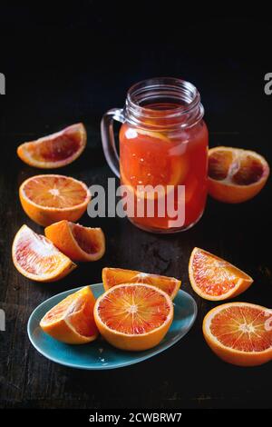 Sizilianische Blutorangen in Scheiben geschnitten in türkisfarbenen Keramikplatte und Glas-Einmachglas von frischen roten Orangensaft über alten Holztisch. Dunkler rustikaler Stil. Stockfoto