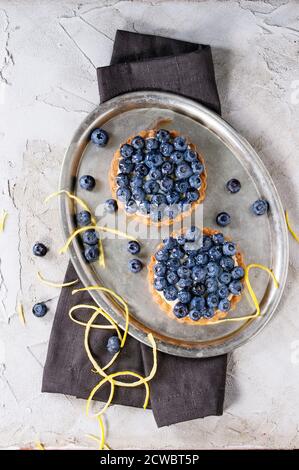 Zwei Lemon Tartlets mit frischen Heidelbeeren, serviert auf Vintage-Metalltablett mit Zitronenschale und schwarzer Textilserviette über grau verputzter Oberfläche. Flach liegend Stockfoto