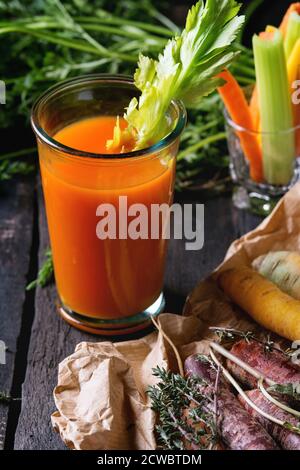 In Scheiben geschnittene bunte rohe Karotten und Sellerie als vegetarischer Snack, und ganze Karotten auf Papiertüte mit einer Glastasse frischer Orangen- und Karottensaft über altem BL Stockfoto