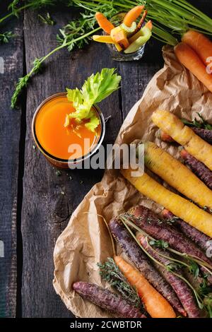 In Scheiben geschnittene bunte rohe Karotten und Sellerie als vegetarischer Snack, und ganze Karotten auf Papiertüte mit einer Glastasse frischer Orangen- und Karottensaft über altem BL Stockfoto