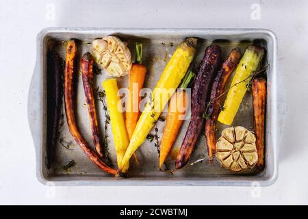 Gebackene ganze bunte Karotten mit Knoblauch und Thymian in Vintage-Aluminium-Tablett über alte Holzoberfläche. Draufsicht Stockfoto