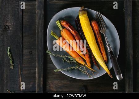 Ganz bunte Karotten mit Thymian in graue Keramik-Platte mit Vintage Gabel über alte Holzoberfläche gebacken. Ansicht von oben Stockfoto