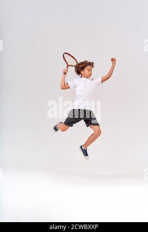 Kleiner Gewinner. Ganzkörperaufnahme eines Jungen im Teenageralter, der mit einem Tennisschläger auf grauem Hintergrund springt, Studioaufnahme. Stockfoto