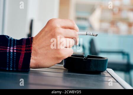 Nahaufnahme eines jungen kaukasischen Mannes, im Pyjama, Rauchen einer Zigarette, die an einem Tisch im Freien auf dem Balkon, in seinem Haus oder in einem Hotel sitzt Stockfoto