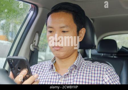 Asian Mann trägt ein kariertes Hemd schaut auf Smartphone in einem Auto. Stockfoto