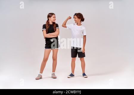 Zwei Jugendliche trainieren zusammen. Boy zeigt seine Bizeps zu einem niedlichen Mädchen, sie stehen isoliert über grauen Hintergrund im Studio Stockfoto