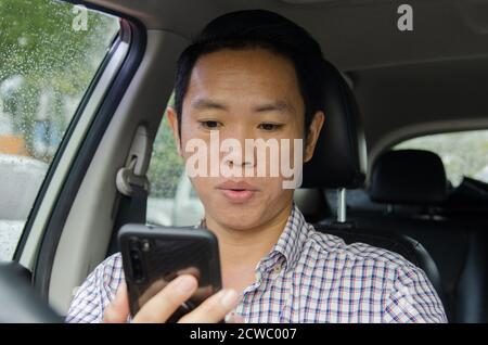 Asian Mann trägt ein kariertes Hemd schaut auf Smartphone in einem Auto. Stockfoto