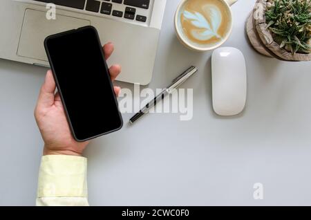 Männliche Hand hält Smartphone, Computer, Stift, Kaffee, Maus auf Schreibtisch Tisch mit Kopieplatz Stockfoto