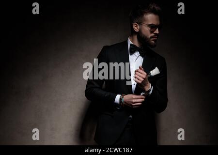 Junger Geschäftsmann trägt eine Sonnenbrille im Stehen und fixiert seine Hülse während Blick beiseite ernst auf dunklen Studio-Hintergrund Stockfoto