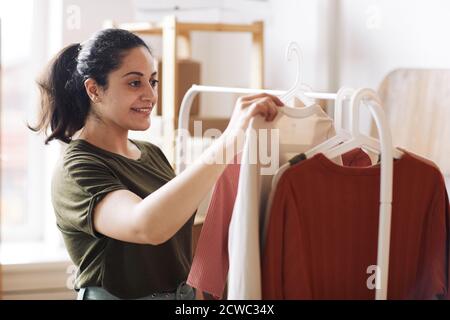 Junge Frau, die in der Nähe des Racks steht und neu anschaut Kleidung, die sie im Bekleidungsgeschäft arbeitet Stockfoto