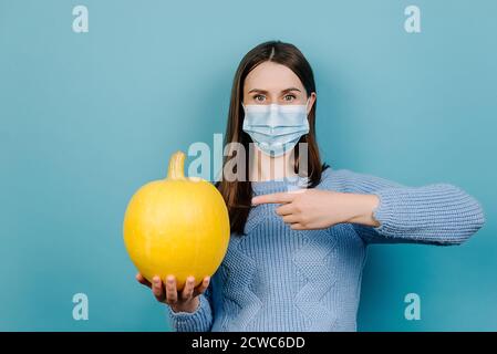 Halloween während covid-19 Pandemiekonzept. Portrait einer jungen Frau mit medizinischer Maske, Zeigefinger auf gelben Kürbis, trägt Pullover Stockfoto