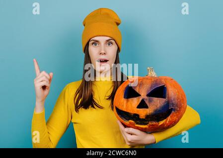 Schockiert junge Frau halten gruselig geschnitzten großen orange Kürbis, zeigt neben auf blauem Studio-Hintergrund mit Kopieplatz, zeigt Ort der Halloween-Party Stockfoto