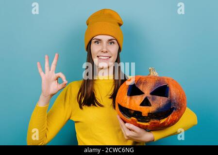 Fröhliche junge Frau zeigt okay Hand Zeichen als ob Genehmigung Vorschlag mit großen gruselig geschnitzten Kürbis, in orange Pullover gekleidet Stockfoto