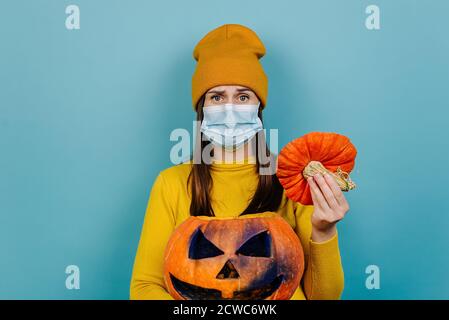 Unglückliche junge Frau trägt medizinische Gesichtsmaske hält gruselig geschnitzten orange Kürbis mit schrecklichen Lächeln, in orange Pullover gekleidet, isoliert auf blauem Studio Stockfoto