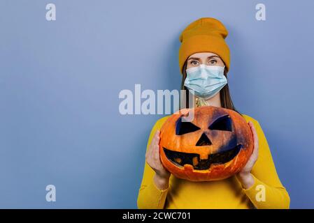 Portrait der jungen Frau trägt schützende medizinische blaue Maske, hält gruselig geschnitzten Kürbis, schaut in die Kamera, in orange Pullover gekleidet Stockfoto