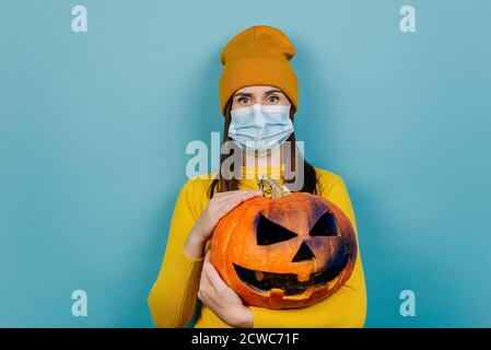 Porträt der tausendjährigen Frau trägt schützende medizinische Maske, hält gruselig geschnitzten Kürbis, in orange Hut und Pullover gekleidet, isoliert über blauem Studio Stockfoto