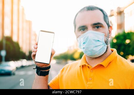 Porträt von 40er Jahren Mann mit chirurgischen medizinischen Maske stehen, halten und zeigt Smartphone-Display. Stockfoto