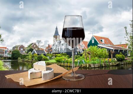 Glas Rotwein und Brie Chhese mit Blick auf Kanal, Brücke und traditionellen hölzernen Fischerhäusern im Dorf Marken, Niederlande. Stockfoto