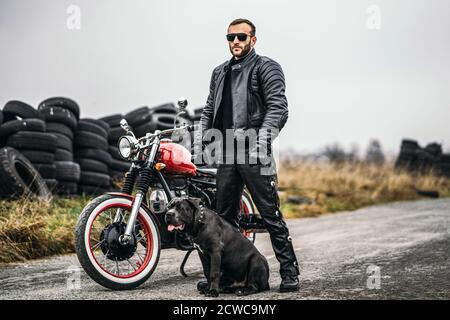 Biker im Lederanzug und sein Hund stehen in der Nähe eines roten Motorrads auf der Straße. Viele Reifen im Hintergrund. Stockfoto