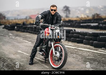 Bärtiger Mann in Sonnenbrille und Lederjacke, der auf die Kamera schaut, während er auf einem Motorrad unterwegs sitzt. Hinter ihm ist eine Reihe von Reifen. Stockfoto