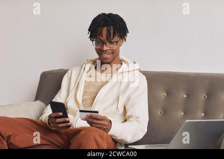 Afrikanischer glücklicher Mann, der auf dem Sofa sitzt und sein Handy benutzt Telefon, um Rechnungen online mit Kreditkarte zu bezahlen Stockfoto