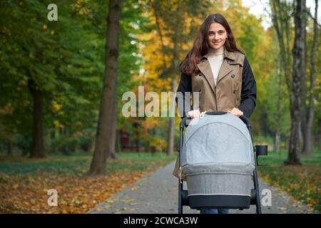 Ansicht von gerade lächelnd Frau, die grauen Kinderwagen in ihren Händen hält. Schönes Mädchen in einem braunen Mantel Blick auf die Kamera und lächelnd. Konzept der Ruhe. Stockfoto