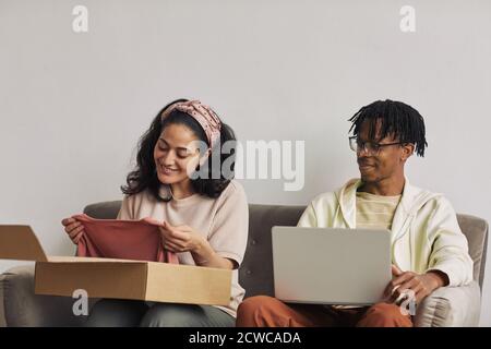 Glückliche junge Frau, die das Paket mit neuem Kleid und Sie mit einem Mann betrachten, der nahe bei ihr sitzt Und mit Laptop Stockfoto