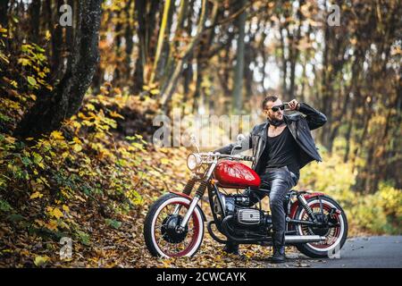 Bärtiger brutaler Mann in Sonnenbrille und Lederjacke sitzt auf einem Motorrad auf der Straße im Wald mit blured bunten Hintergrund. Stockfoto