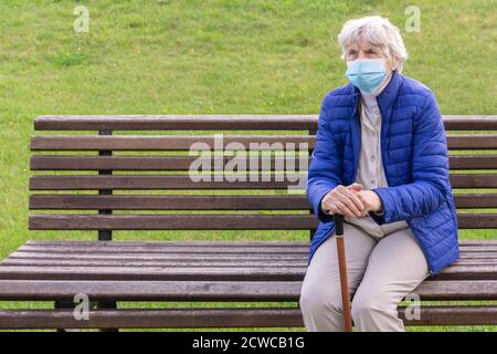 Ältere Frau trägt medizinische Maske sitzt auf der Bank im Freien und hält Spazierstock. Grauhaarige Frau mit schützender Gesichtsmaske auf Stock auf Bank Stockfoto