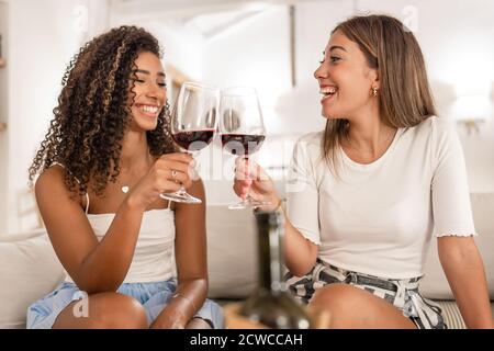 Zwei schöne junge Frauen, hispanische und kaukasische, sitzen auf einem Sofa toasting mit einem Glas Rotwein - multiethnische glückliche weibliche Freunde feiern Stockfoto