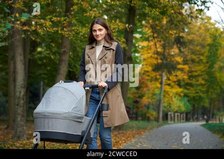 Seitenansicht der jungen Eltern in moderner Kleidung mit einem Kinderwagen in den Händen. Frau, die mit Neugeborenen im grünen Park spazieren geht. Konzept des Parks. Stockfoto