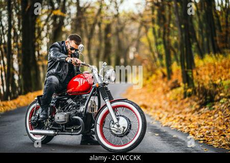 Bärtig brutaler Mann in Sonnenbrille und Lederjacke sitzt auf einem Motorrad auf der Straße im Wald mit verschwommenem bunten Hintergrund. Stockfoto