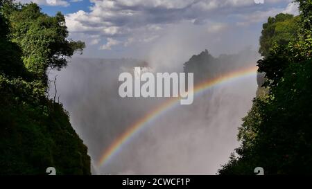 Atemberaubende Aussicht auf den mächtigen Zambesi-Fluss, der aus westlicher Richtung die Victoria Falls hinunter rauscht, mit enormem Sprühnebel und schönem farbigen Regenbogen. Stockfoto