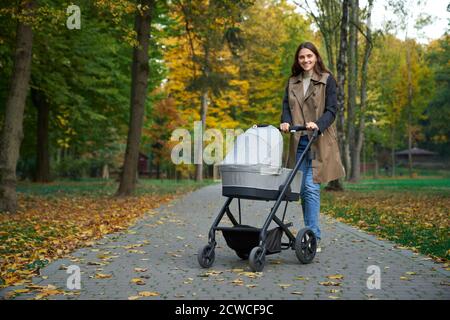 Seitenansicht einer Frau im langen Mantel, die mit einem Kinderwagen im Herbstpark steht. Mutter mit Baby lächelt an der Kamera. Konzept des Kinderwagens. Stockfoto