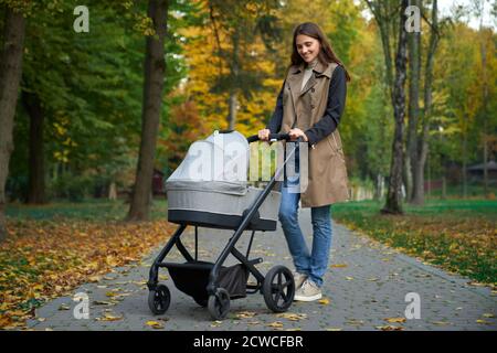 Seitenansicht einer schlanken Frau in voller Höhe im Mantel mit grauem Kinderwagen. Mädchen steht im Park und schaut auf das Baby in grauen Trolley. Konzept des Posing. Stockfoto