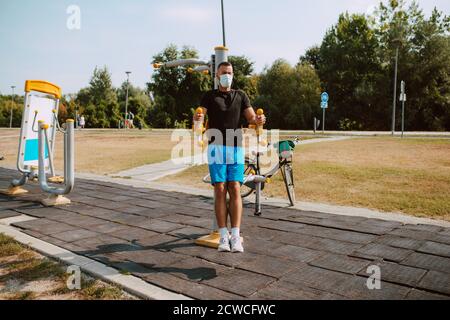 Hübscher junger kaukasischer Athlet mit Gesichtsmaske trainiert auf Arm- und Beingeräten in einem Outdoor-Fitnessstudio. Muskulöser Sportler im Freien. COVID – 19 Stockfoto