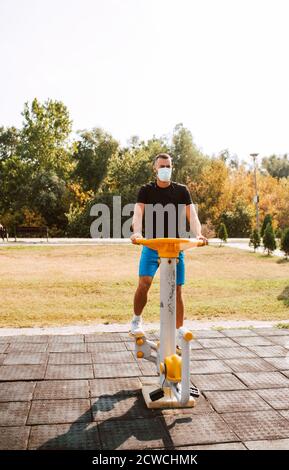 Hübscher junger kaukasischer Athlet mit Gesichtsmaske trainiert auf Arm- und Beingeräten in einem Outdoor-Fitnessstudio. Muskulöser Sportler im Freien. COVID – 19 Stockfoto