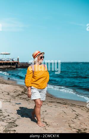 Erwachsene blonde Frau mit Strohsommerhut und gelber Baumwolle shirt Spaziergänge am Strand entlang an einem sonnigen Tag Vertikal Fotoausrichtung Stockfoto