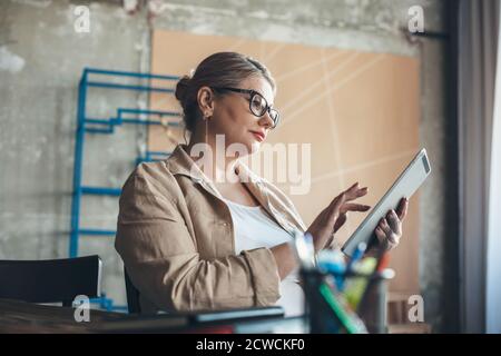 Kaukasische ältere Frau mit blonden Haaren, die auf einer Tablette arbeiten Von zu Hause tragen Brillen und mit einigen Bürosachen Stockfoto