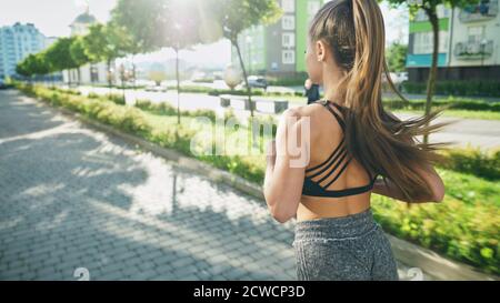 Rückansicht einer nicht erkennbaren muskulösen Frau, die Shorts trägt und in den Straßen der Stadt am Morgen läuft. Fit weibliche Läuferin mit langen Pferdeschwanz üben Cardio-Training im Freien, mehrstöckige Gebäude herum. Stockfoto