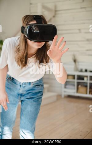 Junge Frau in Jeans und weißem T-Shirt trägt Virtual-Reality-Helm spielt Spiel, arbeitet, schaut Video im Zimmer neben dem Bett. Konzept der modernen Technik Stockfoto